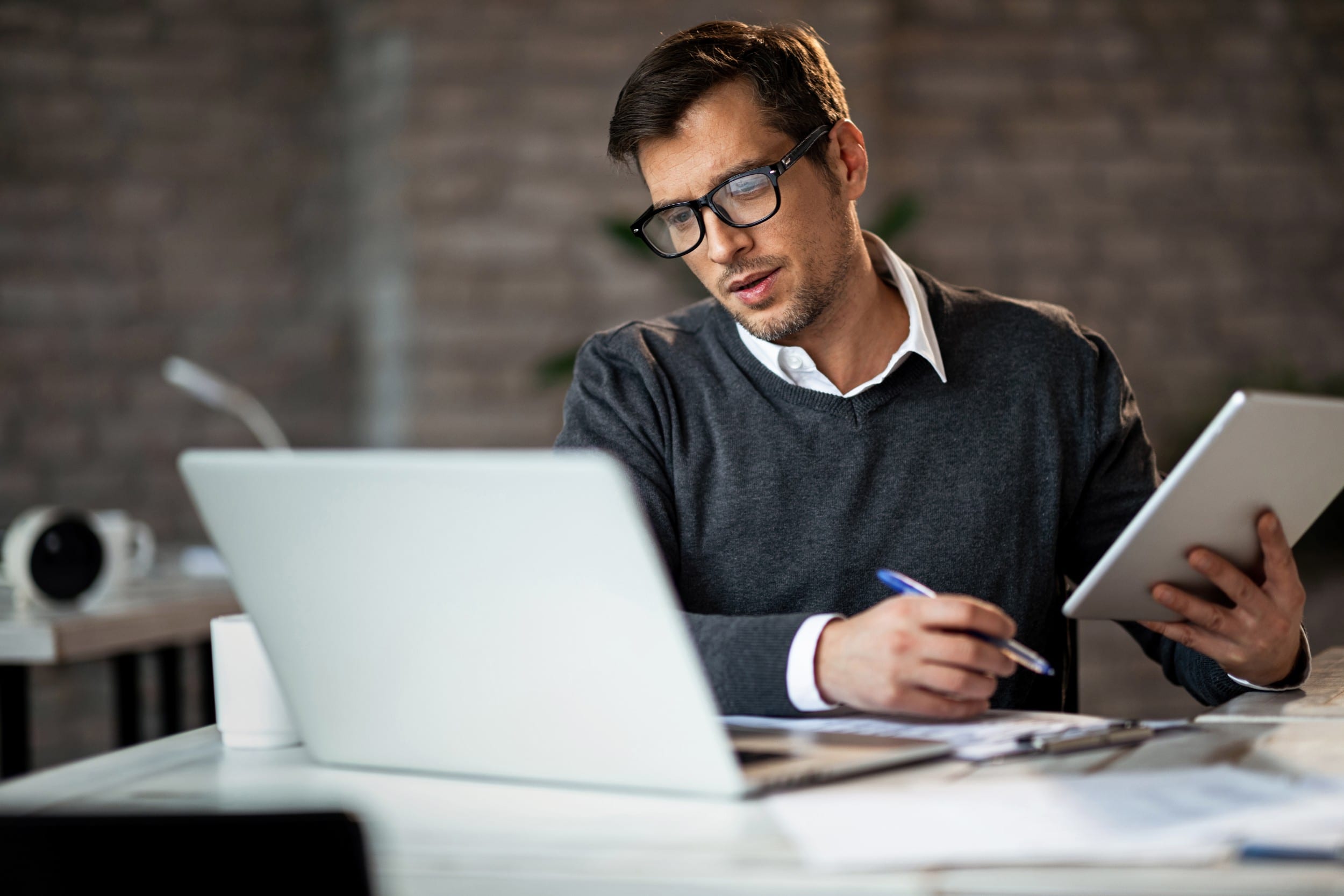 image of man holding pen and tablet whilst looking inquisitively at laptop screen