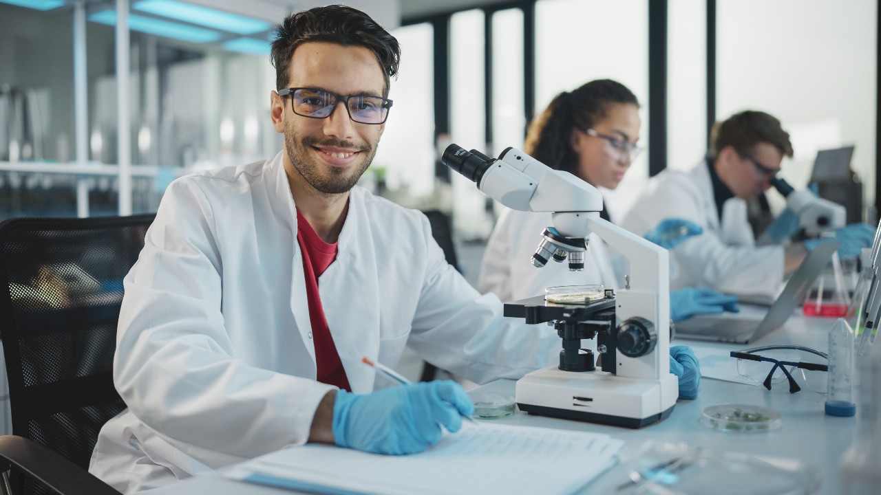 man in a lab coat with a microscope in front of him as he sits next to other people in front of microscopes